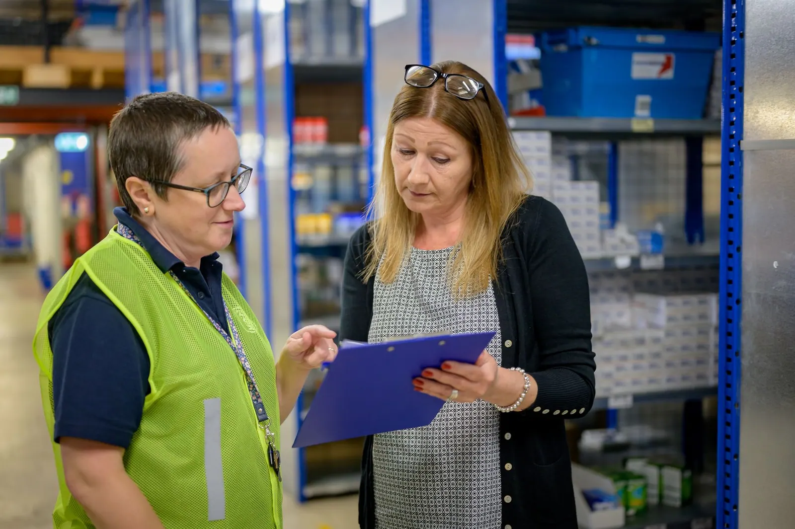 Two women in a warehouse reviewing an inventory checklist on a clipboard.