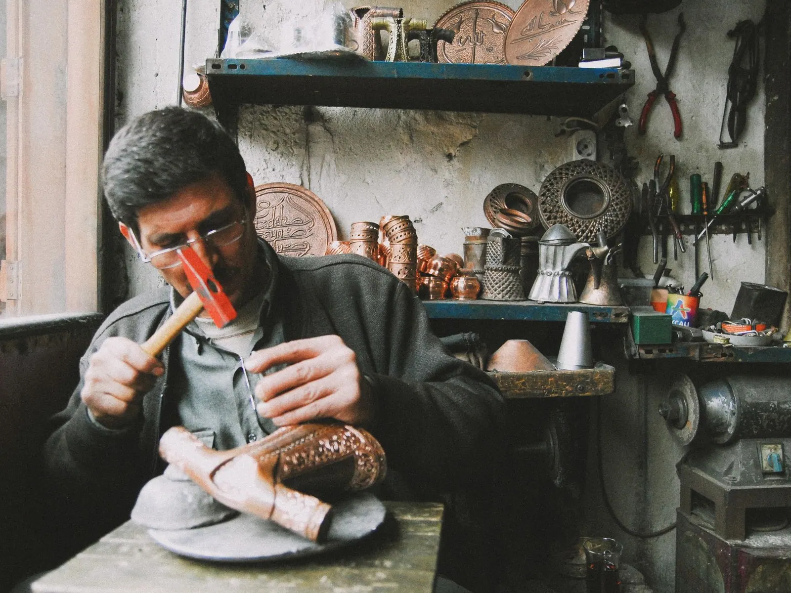 A skilled artisan sculpting copperware with a hammer in a traditional, tool-filled workshop.