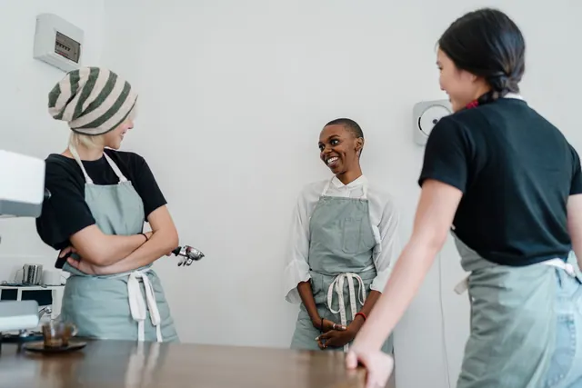 Group of young cheerful diverse female colleagues in aprons smiling and talking to each other while working together in cozy cafe