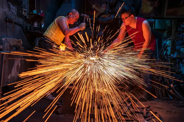 Blacksmiths in Taiwan handcraft metal with sparks flying in an industrial workshop.