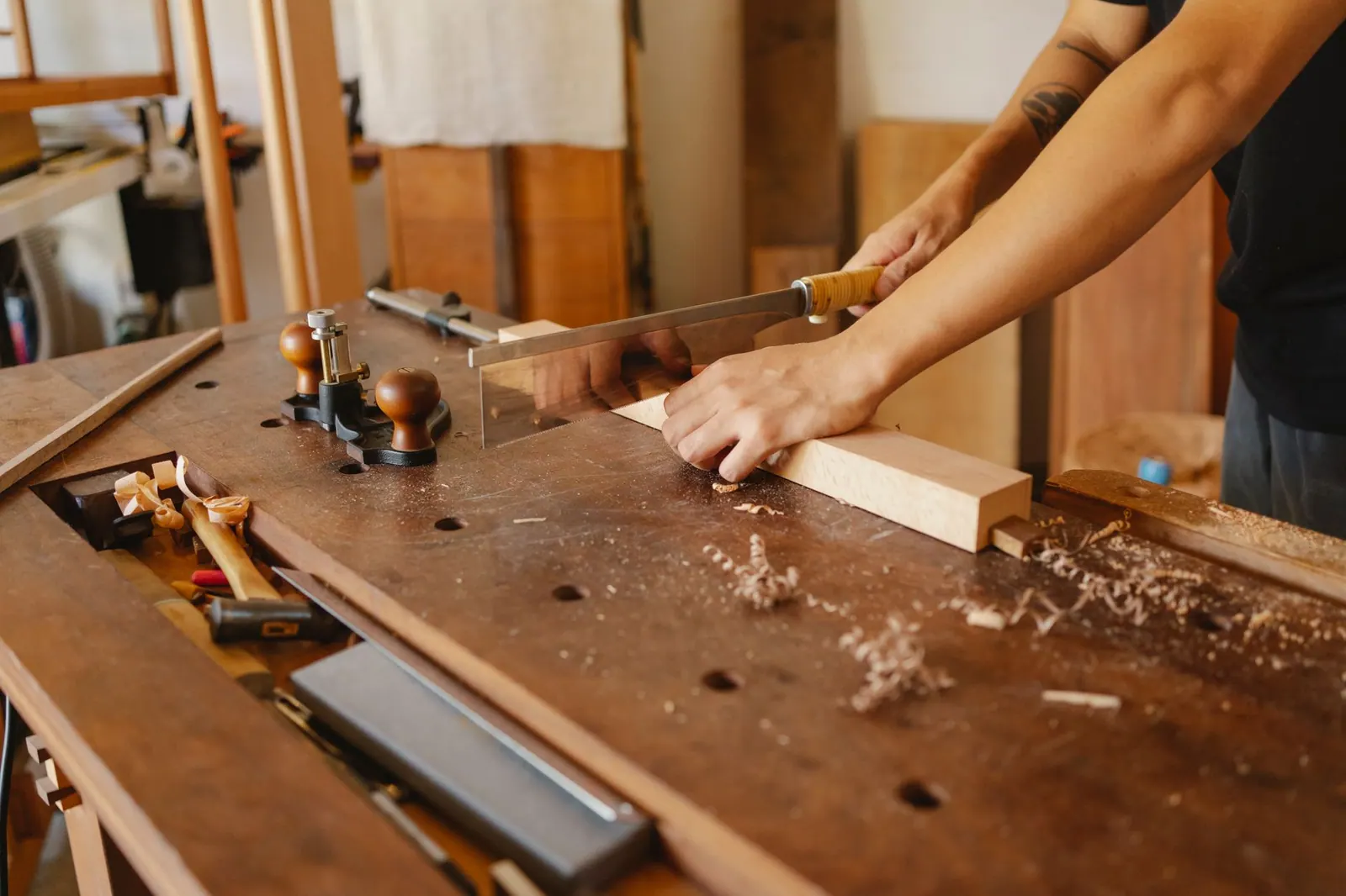 Crop anonymous craftsman with tattoo sawing wooden board with hacksaw at workplace in carpentry workshop