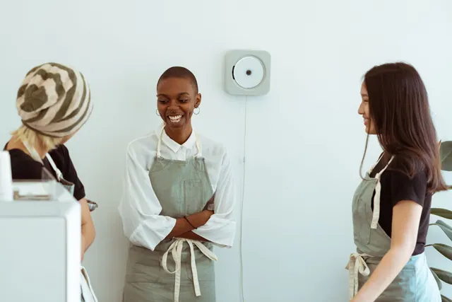Young multiethnic female cafe coworkers in identical aprons talking and laughing cheerfully while having funny daily conversation during break at work