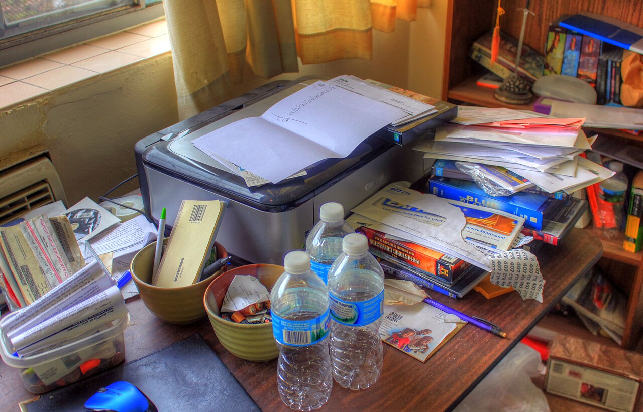 Unordentlicher Büroschreibtisch mit Drucker, Papierstapeln und Büchern