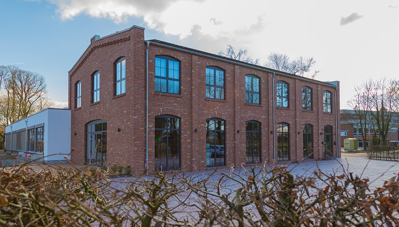 Zweigeschossiges Backsteingebäude mit großen Bogenfenstern und hellem Anbau, davor kahle Sträucher und Bäume.