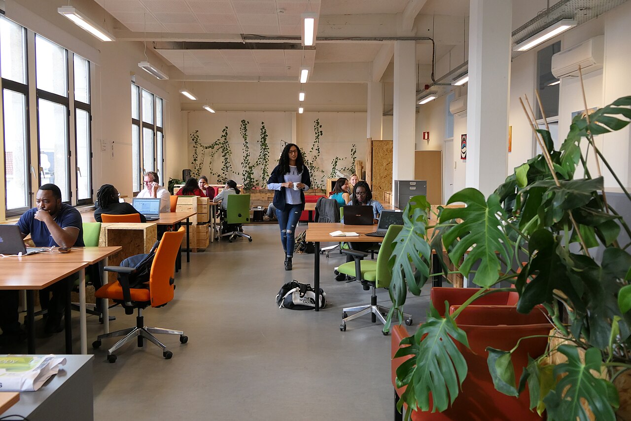 Offener Coworking-Büroraum mit mehreren Personen an Tischen mit Laptops, einer stehenden Person im Mittelgang, großen Fenstern und Pflanzen.
