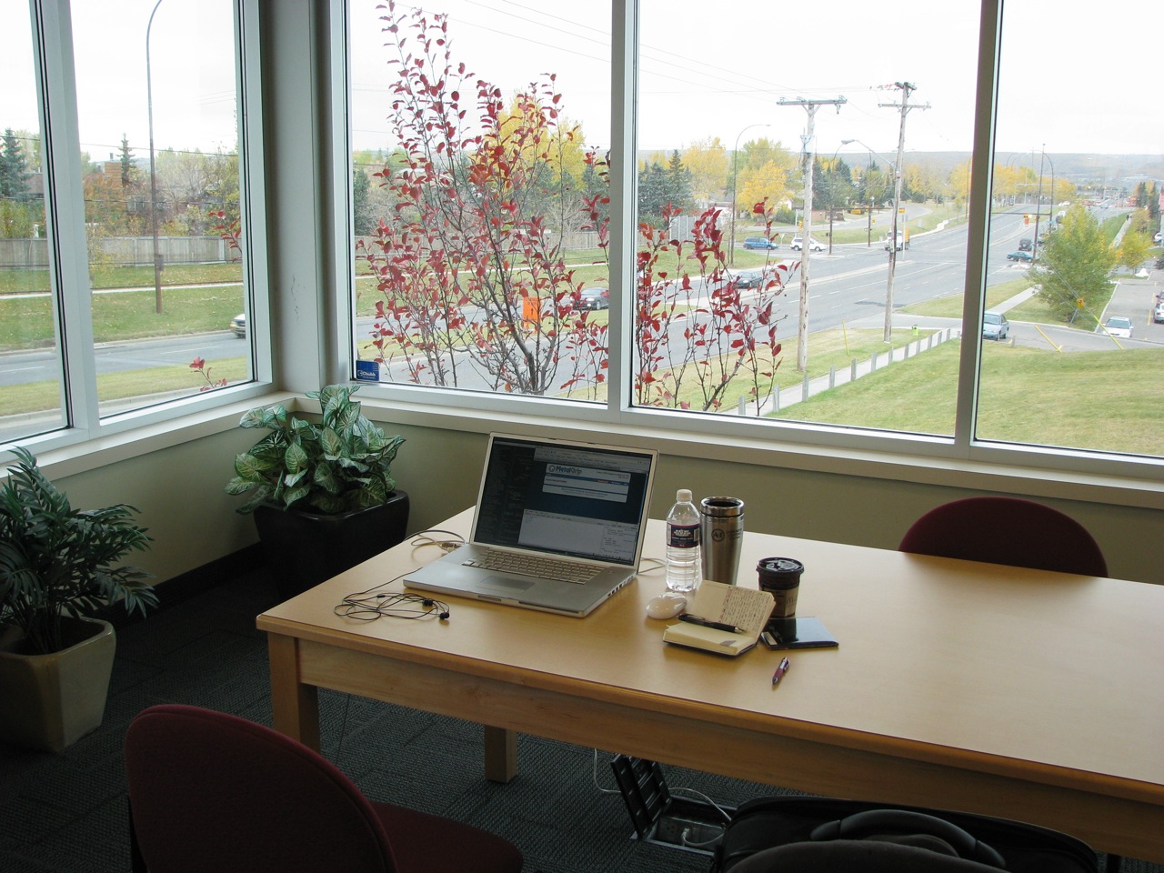 Arbeitsplatz mit Laptop, Notizblock und Getränken vor einem Fenster mit Blick auf herbstliche Bäume