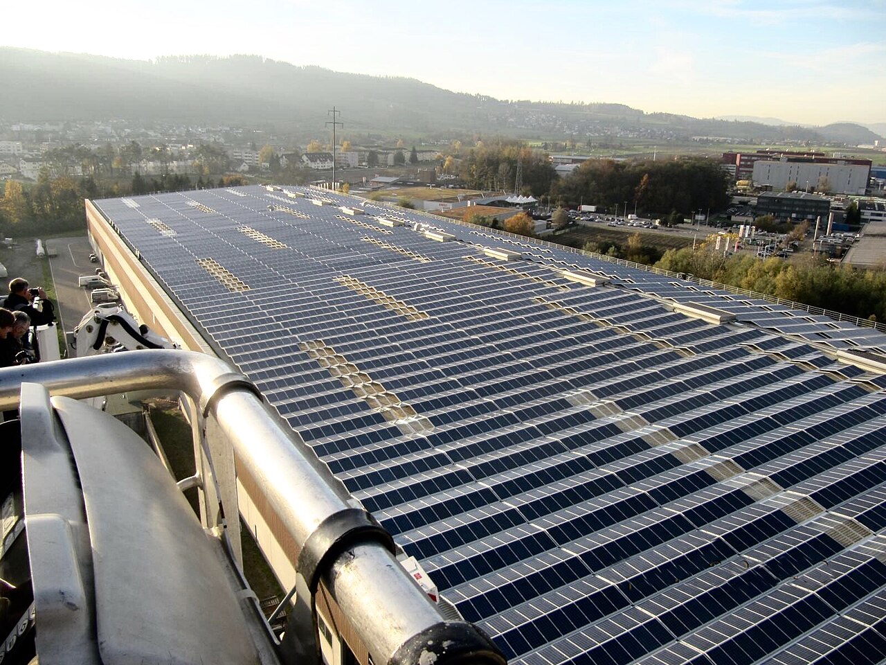 Dach mit dunkelblauen Solarmodulen, im Vordergrund ein Geländer, im Hintergrund Siedlung und Hügel