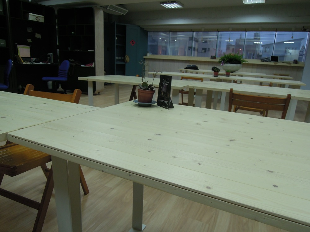 Coworking-Bereich mit langen Holztischen, Stühlen, Pflanze und Fensterfront bei Dämmerung.