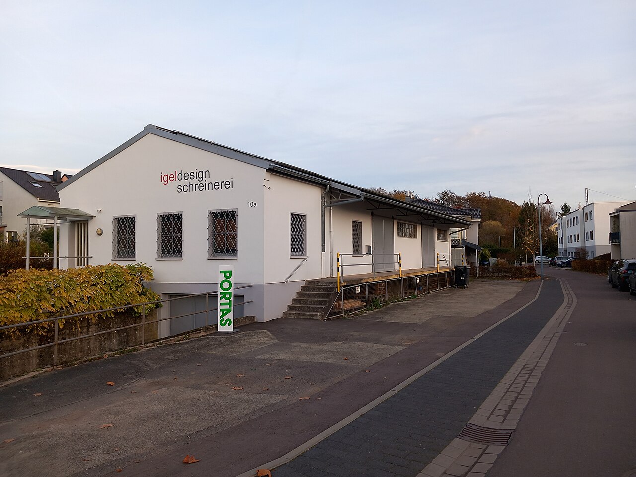 Außenansicht der Schreinerei Igeldesign mit weißem Gebäude, roter Firmenbeschriftung und grünem Portis-Schild.
