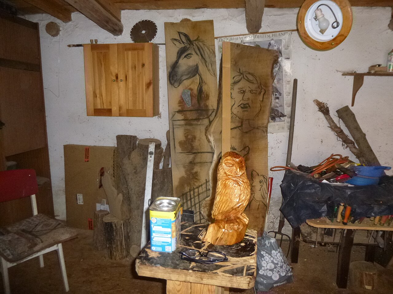 Werkstatt mit geschnitzter Holzeule, Holztafeln mit Pferdekopf und Gesicht, und Werkzeug auf einem Tisch