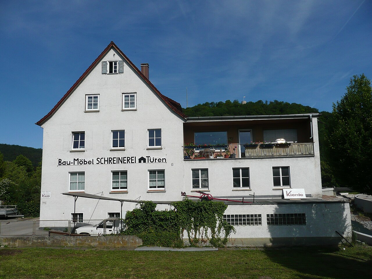 Mehrstöckiges weißes Gebäude einer Schreinerei mit Satteldach, schwarzer Beschriftung 'Bau-Möbel SCHREINEREI' und Anbau mit Balkon