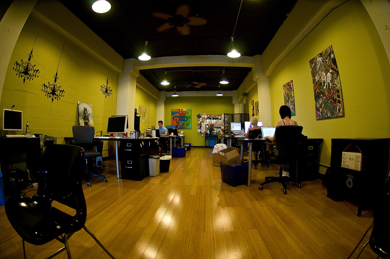 Coworking-Büro mit gelben Wänden, Holzfußboden, Schreibtischen und zwei Personen an Computern.